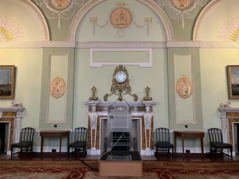 The court room in the Bank of England which has decorated green walls, a gold clock above a fireplace and a patterned rug. A lectern with Bank of England written on it stands in the middle of the room 