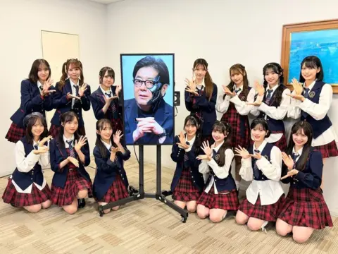 AKB48 Members of AKB48 pose next to a screen showing an "AI" image of their composer Yasushi Akimoto