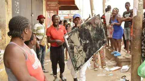 AFP via Getty Images A man in a baseball cap carries a large-screen television as he walks down a street in Luanda. There are people lining the pavement looking at him.