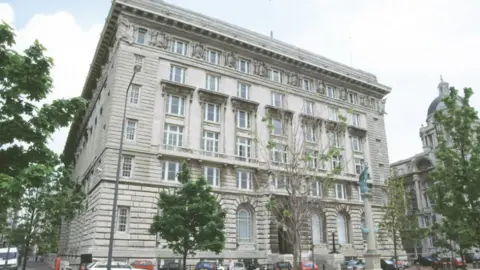 An exterior shot of the early 20th century-built Cunard Building on the Liverpool waterfront. It is an off-white six-storey Portland stone building built in a mixture of Italian and Greek renaissance styles and is surrounded by young trees.