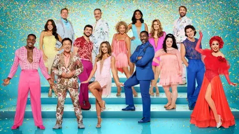 The 15 cast members of this year's Strictly Come Dancing in colourful, sparkly and frilly outfits. Some of them are standing on steps behind the others and there is gold glitter falling on them.