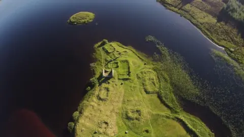 Open Virtual Worlds at University of St Andrews A drone image showing archaeological remains at Finlaggan. There are ruined walls and rectangular shapes beneath grass-covered ground on an area of land that sticks out into the loch. The loch's waters are a peaty brown.