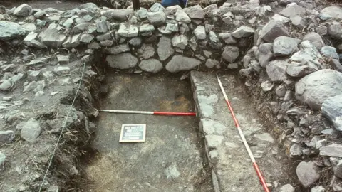 Dr David Caldwell An exposed area of stone-built walls and a floor. There are red and white measuring poles laid out on the ground. 