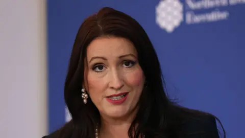 PA Emma Little-Pengelly has long black hair and is wearing makeup, including pink lipstick. She has a pearl necklace and earrings and is wearing a black blazer.