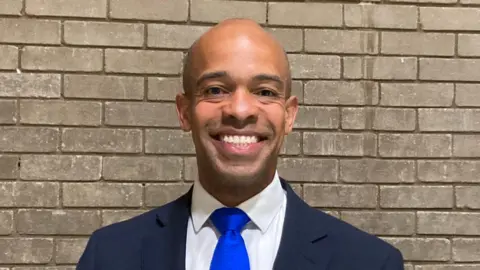 Ben Obese-Jecty is smiling in front of a grey brick wall. He is wearing a dark blazer, white shirt and bright blue tie.