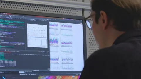 Jeff Dowling/EMBL-EBI The back of a dark-haired man's head and shoulders as he looks at a computer screen. You can see lines of computer code and multicoloured graphs being produced although their meaning is unclear form the image.