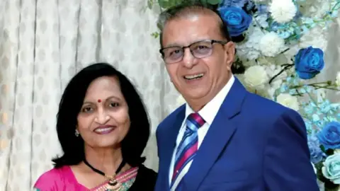 Miten Patel Shobhana Patel and Ashok Patel smile for the camera in front of a patterned white curtain and a blue and white floral display. Shobhana has short black hair and wears a bright pink top, while Ashok has short black hair and wears a blue suit jacket, a white shirt and a pink and blue striped tie.