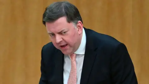 Getty Images Colin Smyth, a man with dark hair, standing in a stooped position in the Scottish Parliament. He is wearing a dark suit, white shirt and pink tie. There is wood panelling in the background. 