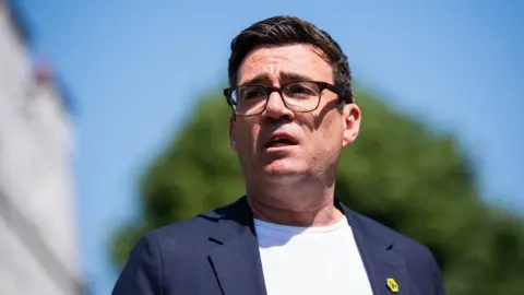 PA Media Greater Manchester Mayor Andy Burnham stands wearing a white t-shirt and a jacket with a yellow bee pin on his lapel. He looks away from the camera.