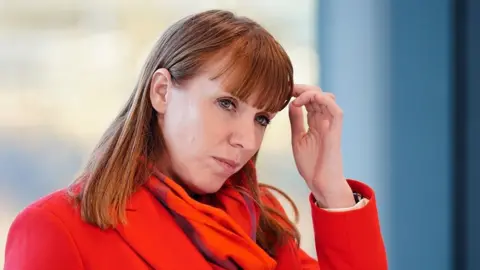 PA Media Angela Rayner wears a thoughtful expression and scratches her head. She is wearing a bright red coat