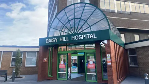 BBC A large brown bricked building has a green banner on it's front that reads 'DAISY HILL HOSPITAL.' The banner sits at the entrance to the building below a glass archway which is encased in a green skeletal structure. They sit on-top of a red brick extension out the front of the brown bricked building. There is a black bench and a small green tree to the left of the entrance