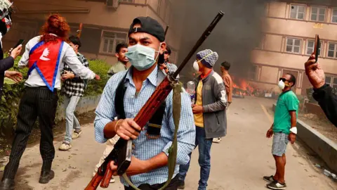 Getty Images A man in a face mask and a backwards cap carries a rifle while others gather behind him amid fire and smoke