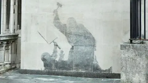 PA Media Banksy mural of a judge attacking a protestor with a gavel. The painting has been washed away, so the details are not visible, leaving a grey shadowy silhouette.