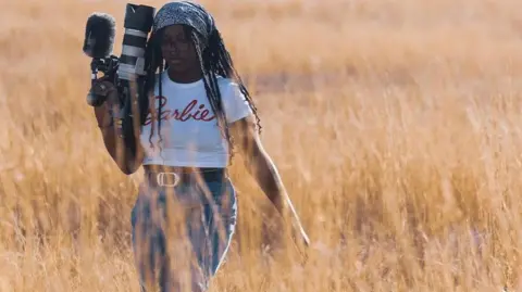 Eshe Powell Eshe Powell in a field with long grass around her, holding a camera. She is wearing a white T-shirt and jeans. She has one arm held out. She has long hair with a headscarf on her head. 