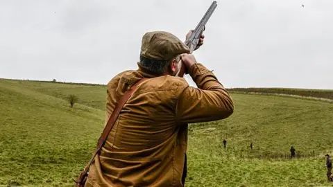Getty Images A man wearing a brown jacket and flat cap aims a gun into the air where there are pheasants flying.  