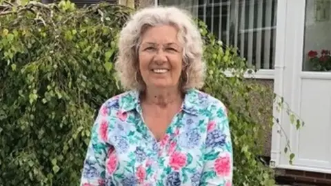 Family handout Valerie Kneale with shoulder-length grey curly hair wearing a pink, lilac and green floral blouse smiling in front of a shrub. She is standing in front of a pebbledashed house with a white plastic door and windows.