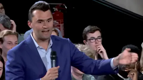 Cambridge Union/YouTube Charlie Kirk in a blue suit and shirt, holding a microphone, with short dark brown hair, gesticulating and talking into a microphone. There are people sitting behind him spectating.
