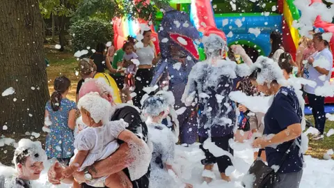 Little Miracles Children and parents playing with white foam- with a colourful bouncy castle in the background.