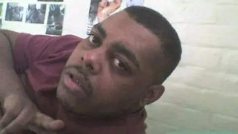 Leroy Douglas A selfie of Leroy. He is pointing at the camera, with his head and shoulders visible. He is wearing a burgundy t-shirt and has stubble and short black hair. A white brick background with posters on the wall can be seen behind him.