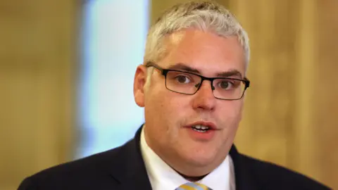 Liam McBurney/PA Wire An archive photo of DUP leader Gavin Robinson speaking to the media at Stormont earlier this year. He has short grey hair and black rimmed-glasses. He is wearing a black suit, a white shirt and a silver and gold stripped tie.