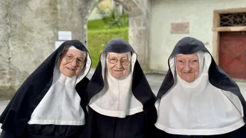 BBC Three nuns stand in front of the monastery in their habits, with Sister Rita on the left and Sister Regina in the centre both wearing glasses