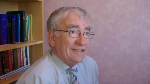 BBC A grey-haired man with glasses, wearing a grey shirt and striped tie, looks beyond the camera. In the background is a bookcase