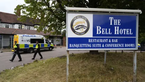 PA Media Two police officers walking next to the hotel, which is surrounded by a thin metal fence. A police van has also parked next to the building. In the foreground is a large blue sign which reads 'The Bell Hotel'.