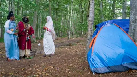 The three members of the tribe, dressed in knitted jumpers and robes with tribal jewellery, stand next to two blue tents in the woodland.