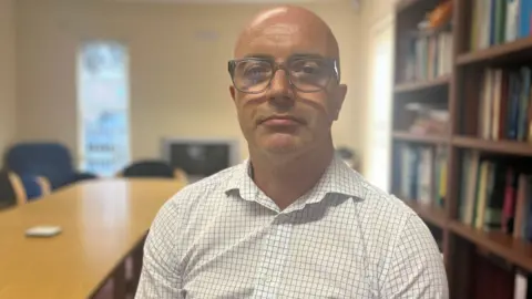 BBC Dr Ciaran Mullan he is wearing a white check shirt and glasses.