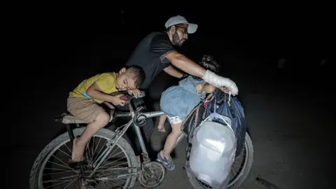 Anadolu via Getty Images A man with a bandaged hand steers the handlebars of a bicycle while two small children lie on the front bars and seat. Some bags hang from the bike's handlebars as they journey in the night, with a bright light shining on to them.