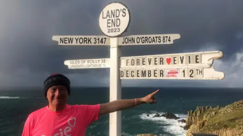 The Evie Dove Foundation An older man wearing a bright pink T-shirt and a black beanie smiles as he points to a sign at Lands End in Cornwall with distance markers to the Isles of Scilly, New York, and John O'Groats. One of the signposts reads "Forever Evie, December 12th".