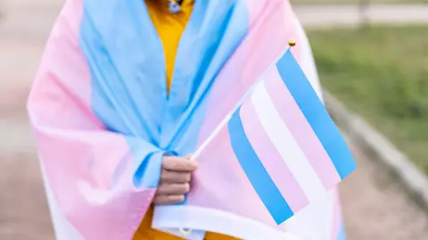 Getty Images A person is draped in the transgender flag. They are also holding a small pink, blue and white flag.