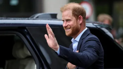 Reuters Prince Harry - a slightly-balding man with red hair and a red beard waves as he steps into a dark coloured car. He wears a pale blue shirt and a navy blue suit, with a brown beaded bracelet.