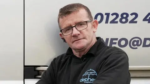 Reuters James Anderson standing in front of his van. He is wearing a black jumper with branding for his company Defer. He has short brown hair and black glasses.