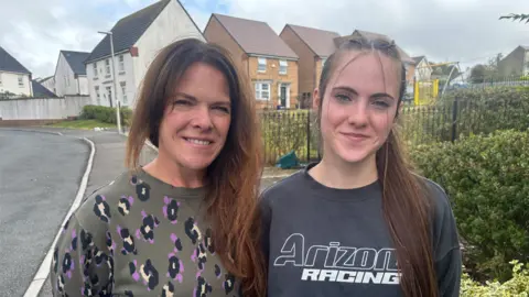 BBC A woman with long brown hair in a green/brown jumper with purple, black and white leopard print, stood next to her daughter who has long brown hair pulled back in a ponytail in a grey/blue jumper with "Arizona racing" written on the front