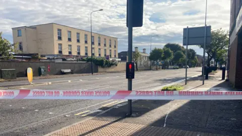 BBC A street with a traffic light and crossing visible. There is a two-storey beige building to the left of the street. Police tape is across the centre of the image.