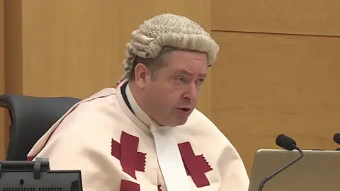 BBC a high court judge in a wig and white robe with red crosses on it sits in court