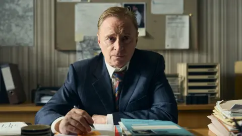 ITV Robert Carlyle in a photo from the Hack. He is sat at a desk surrounded by bundles of paperwork. He wears a dark blue suit and a white shirt with the top button undone and a tie that has been loosened. He wears a tired and worn expression on his face and looks contemplative. He is holding a pen but does not look ready to write.