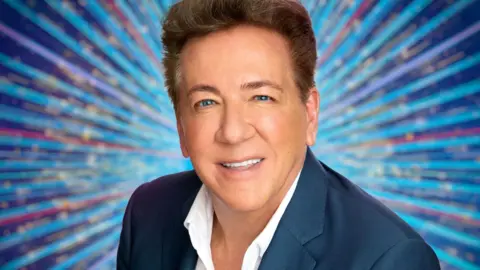 BBC Ross King is smiling at the camera. He has short, dark hair and is wearing a white shirt and blue suit jacket. The background is different shades of blue and red lines with gold sparkles.