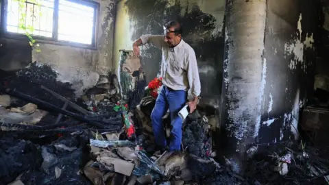 BBC A man in a pale shirt and jeans stands in a room blackened by fire holding burnt items including colourful artificial flowers.