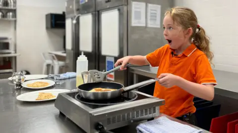 BBC Ophelia is in an orange shirt cooking pancakes with a spatula in a commercial kitchen, surrounded by stainless steel appliances, metal bowls, and plates of pancakes on the counter