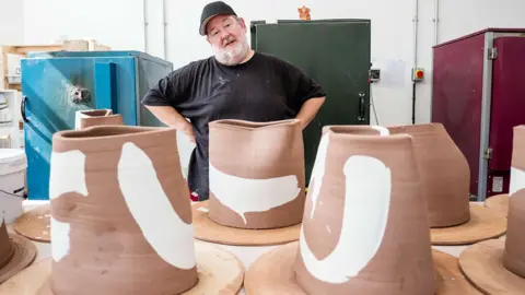 British Ceramics Biennial Johnny Vegas stands in a workshop with his hands on his hips and head cocked to one side. He is standing behind a table with large brown clay models with white paint on them.
