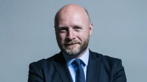 UK Parliament A portrait style photo of Liam Byrne, who is wearing a dark suit, pale blue shirt and navy tie, posed against a grey backdrop. He has a beard and balding head.