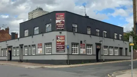 Graham Matthews A grey building with a darker grey roof, it has signs on and is on the street corner