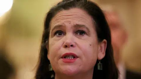 PA A woman with red lipstick on, green dangly earrings and black hair and brown eyes looking into the distance mid talking with a blurred background