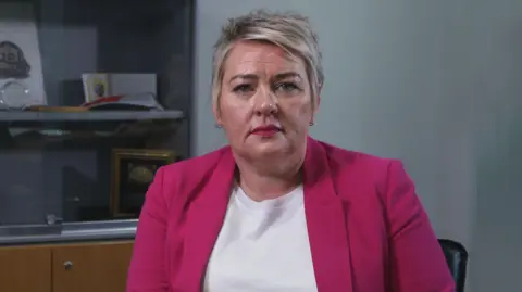 BBC A brown-eyed woman with short cut blonde hair looks directly at the camera. She is wearing a white t-shirt and pink blazer, with two gold hooped earrings in. The woman is sitting in a black chair in a dimly lit grey office. Behind her is a cabinet with a glass door, showing policing plaques and memorabilia.