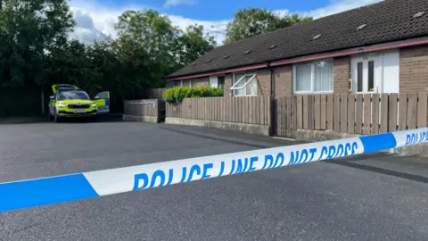 BBC Blue and white police tape in front of a house. In the background, there is a PSNI car. Behind the car, there are some trees.
