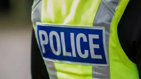 BBC The back of a police officer wearing a hi-vis yellow jacket with the word "police" written in write on a blue background