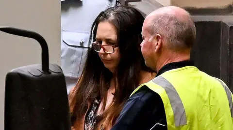 Getty Images A woman with long brown hair, wearing glasses, stands between a vehicle and a man in a yellow high-vis vest