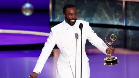 Christopher Polk/Variety via Getty Images Tramell Tillman smiles while holding an Emmy award onstage, wearing a white bowtie, shirt and suit, as he wins the outstanding supporting actor in a drama series award at the Emmys.